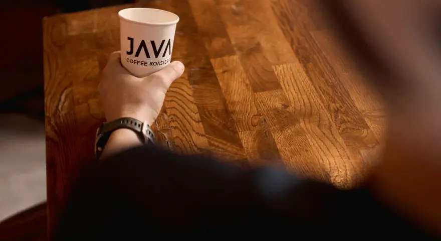 Dłoń trzyma kubek z logo "JAVA COFFEE ROASTERS", gotowa do degustacji świeżego batch brew na drewnianym stole.