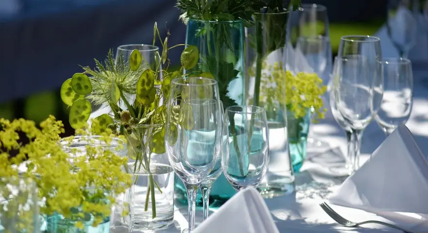 Eleganckie wesele w ogrodzie. Stół nakryty białym obrusem, ozdobiony zielonymi roślinami i kieliszkami.