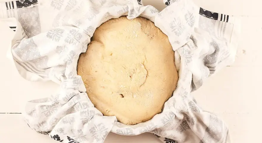 Masa de pan de patata cruda, cubierta con un paño blanco con estampado floral gris, lista para levar.