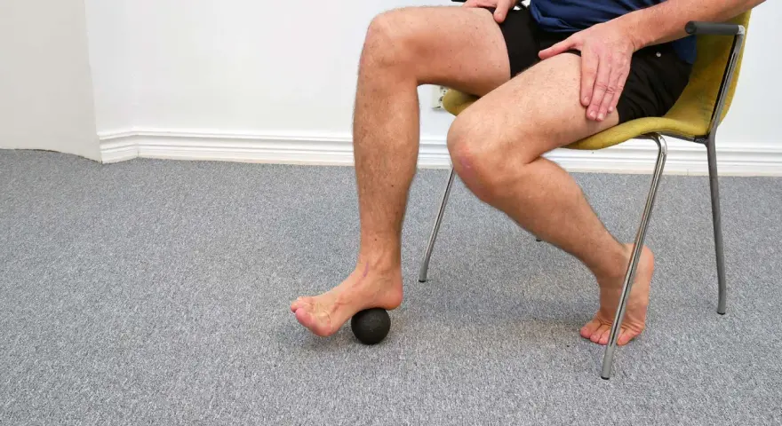 Man sitter på en stol och använder en liten boll för att rulla under foten.