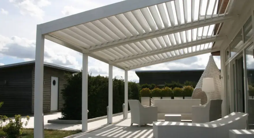 Un moderno porche de obra con techo de lamas ajustables, mobiliario de exterior blanco y un camino de madera que conduce a una cabaña.