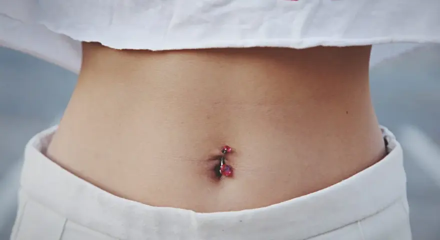 Brzuszek z kolczykiem w pępku. Czy dziurka w pępku zarasta? Delikatna skóra, białe spodenki i crop top z czerwonym napisem.