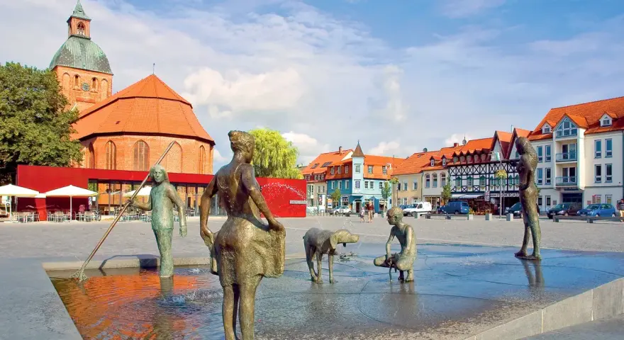 Bronzefiguren am Brunnen in Ribnitz-Damgarten, mit Kirche und Fachwerkhäusern im Hintergrund. Ein Ort, der an Bernstein erinnert.