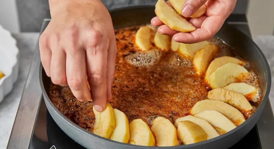 Hände legen Apfelschnitze in eine Pfanne mit karamellisierter Butter. Die Zubereitung der tarte tatin original beginnt.