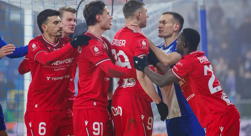 Piłkarze w czerwonych strojach kłócą się z zawodnikiem w niebieskiej koszulce.