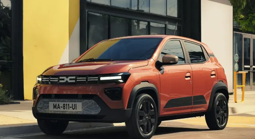 Pomarańczowy Dacia Spring stoi przed budynkiem z żółtą ścianą.