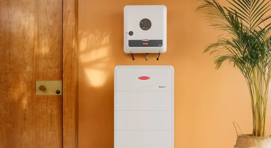 System magazynowania energii Fronius Symo GEN24 i akumulator Reserva na pomarańczowej ścianie, obok drewnianych drzwi i rośliny.