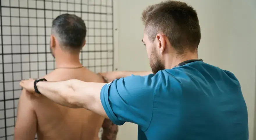 Therapeut führt krankengymnastik schulter Übungen durch. Ein Mann steht vor einem Gitter, während ein Therapeut seine Schultern berührt.