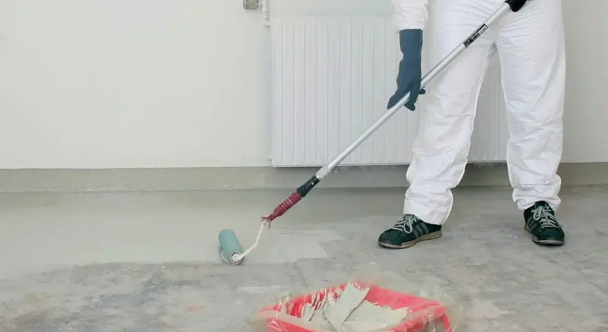 Person i skyddsdräkt målar betonggolv med en roller. Färgen är ljus och en färgtråg syns på golvet.