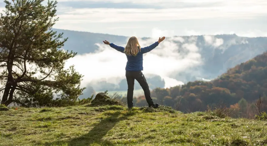 Frau mit ausgebreiteten Armen auf einem Hügel, umgeben von Nebel und Herbstfarben. Das Gefühl von Freiheit und die Bedeutung des Pilgerns werden hier spürbar.