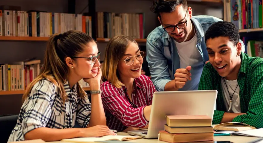 Vier Studierende lernen in der Bibliothek. Ein Mann, der wie ein seelsorger quereinsteiger wirkt, erklärt etwas am Laptop.