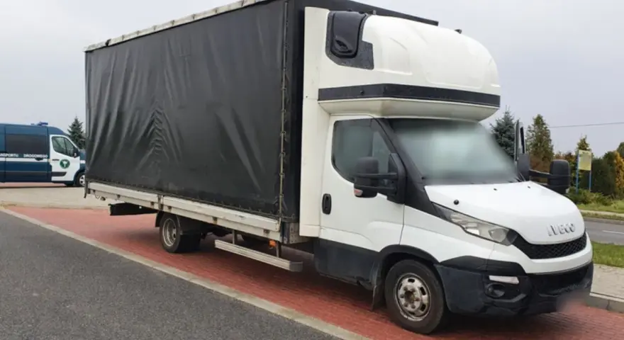 Biały bus Iveco z czarnym plandeką, ładowność 1,5 tony, stoi na czerwonej kostce brukowej.