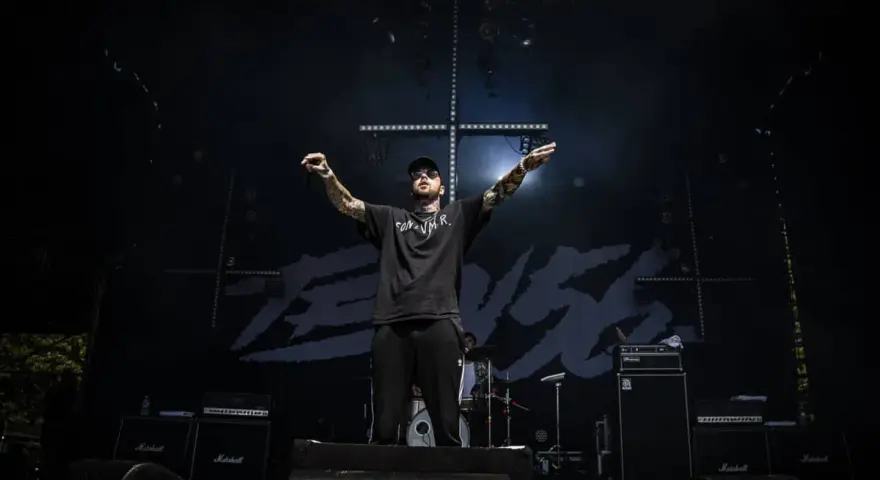 Un rappeur tatoué, casquette et lunettes de soleil, bras levés sur scène, devant un fond noir avec le logo "TEN 56".