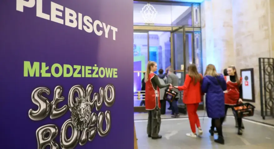 Plakat "PLEBISCYT Młodzieżowe Słowo Roku" z lwem w koronie. W tle młodzież wchodzi do budynku.