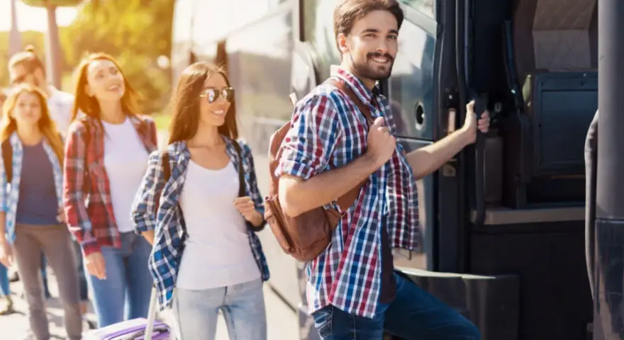 Grupa młodych ludzi z plecakami i walizką wsiada do autobusu, gotowa na wycieczki fakultatywne.