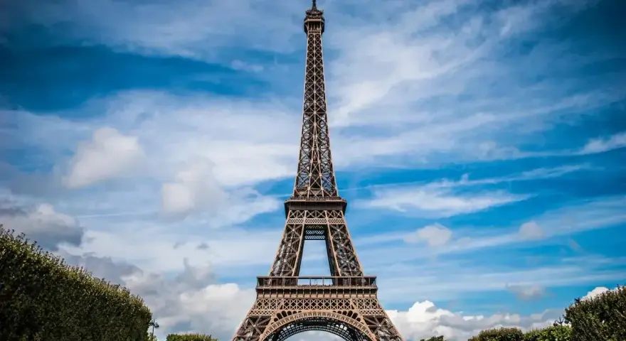 Majestatyczna wieża Eiffel w Paryżu, górująca nad zielonymi krzewami i błękitnym niebem z chmurami.
