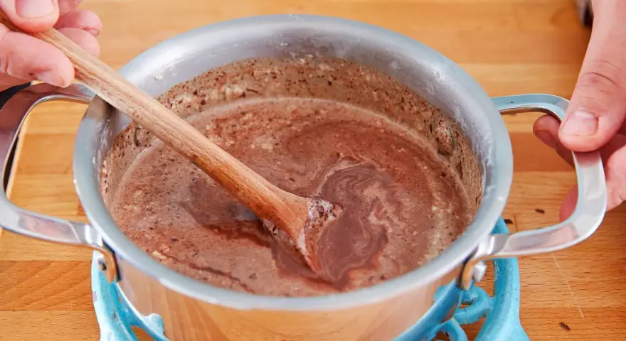 Hände rühren eine schokoladige Masse in einem Topf. So einfach lässt sich die Glasur selber machen für Kuchen oder Gebäck.
