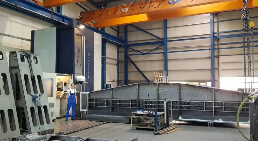 Ein junger Zerspanungsmechaniker in blauer Latzhose steht vor einer großen Soraluce-Maschine in einer Werkshalle. Daneben liegen massive Metallteile.