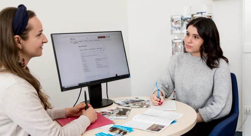 Zwei Frauen besprechen am Tisch eine Webseite zur allgemeinen Studienberatung. Eine Frau schreibt Notizen, die andere schaut auf den Monitor.
