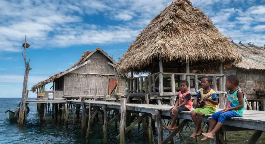 Trzech chłopców siedzi na drewnianym pomoście przy tradycyjnych chatach na wodzie w Raja Ampat.
