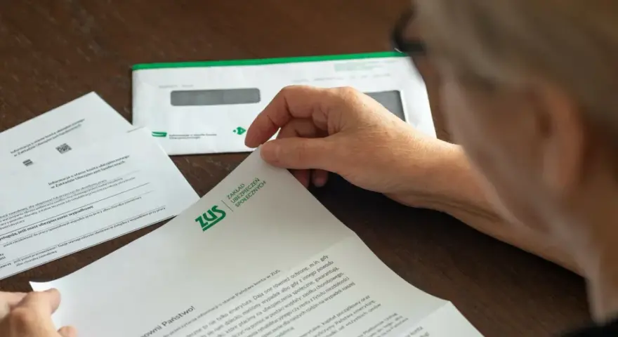 Dłoń starszej osoby przegląda dokumenty z ZUS. W tle koperta z logo ZUS.