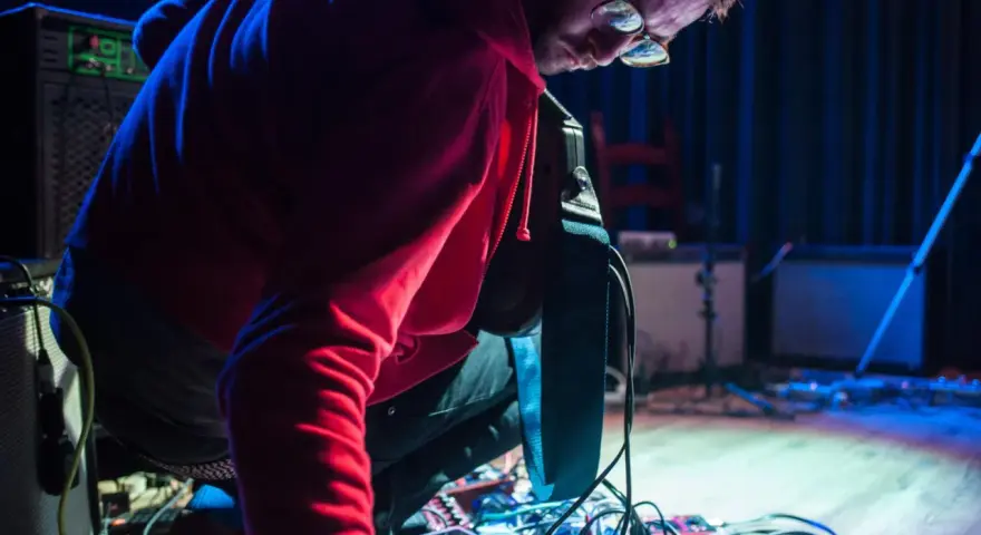 Musiker mit roten Kapuzenpullover beugt sich über ein Pedalboard, um den "Delay"-Effekt einzustellen.