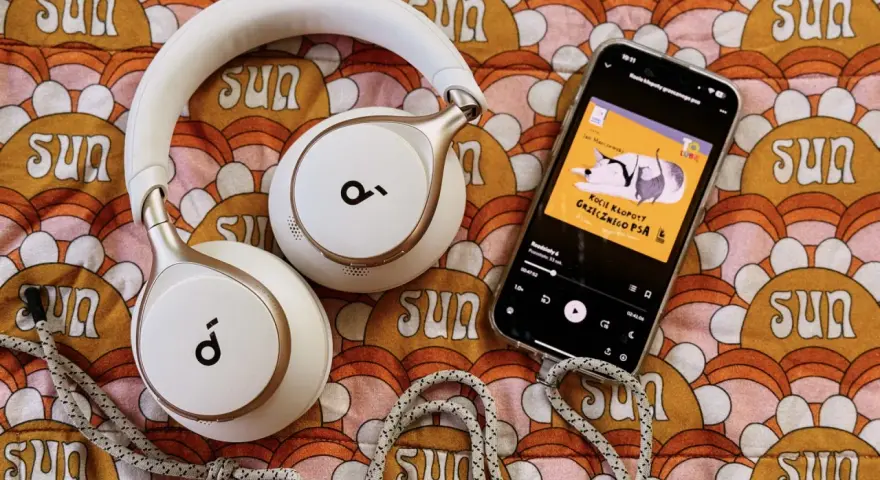 Białe słuchawki i smartfon z okładką audiobooka "Kocie kłopoty grzecznego psa" leżą na kolorowym tle z motywem słońc. Idealne do słuchania książek z lektorem.