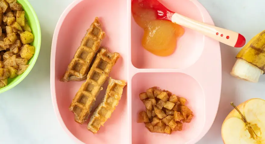 Baby-freundliche Apfelwaffeln mit Apfelmus und Zimt-Apfelstückchen auf einem rosa Teller. Ein halber Apfel und eine Banane liegen daneben.