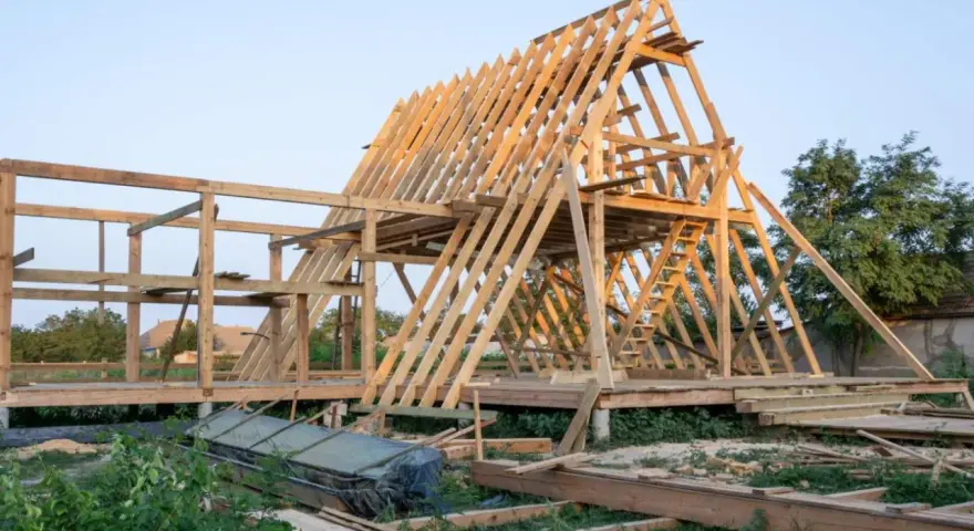 Drewniana konstrukcja domu w budowie. Widoczny szkielet dachu i ścian, schody. Fundament punktowy pod domek drewniany jest już gotowy.
