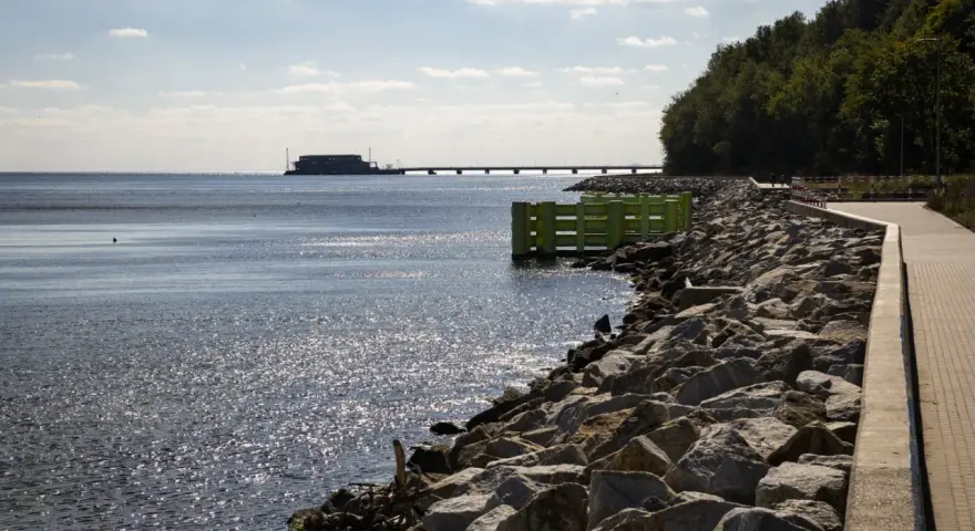 Kamieniste wybrzeże w Gdyni Oksywie z zielonym pomostem i falochronem. W oddali widać most i budynek na wodzie.
