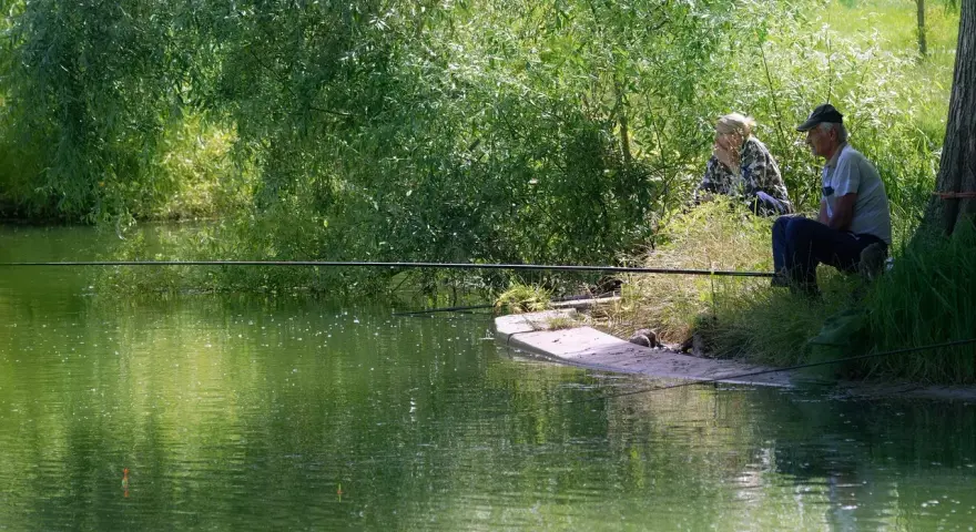 Dwaj wędkarze siedzą nad zieloną wodą, czekając na branie. Jeden z nich, starszy mężczyzna, skupiony na swoim sprzęcie, gotowy do **casting spinning**.