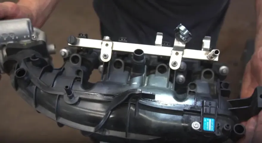 Hands hold a black plastic intake manifold with a fuel rail and sensors. This part is crucial for intake manifold runner control.