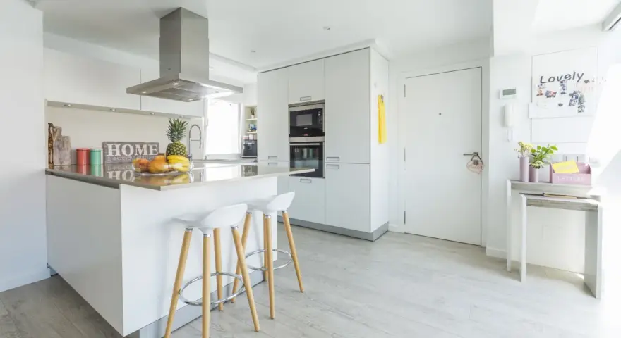 Moderna cocina blanca y gris claro con isla central, taburetes y frutas frescas.