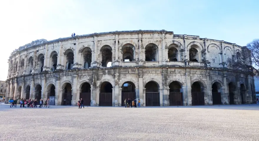 The ancient Roman amphitheater in Nîmes, France, stands majestically under a clear blue sky. Tourists gather in the plaza, ready to explore this historic marvel.