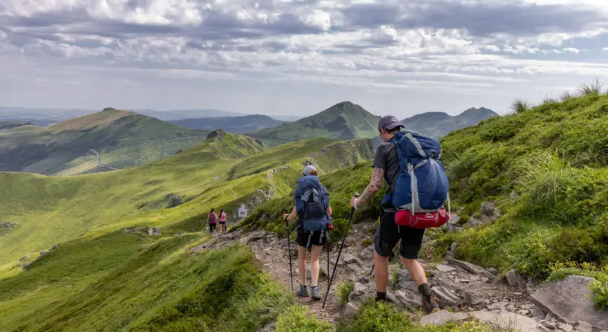 Randonnée sur le Puy Mary, des marcheurs explorent un sentier rocailleux bordé de verdure, avec des montagnes ondulantes en arrière-plan sous un ciel nuageux.