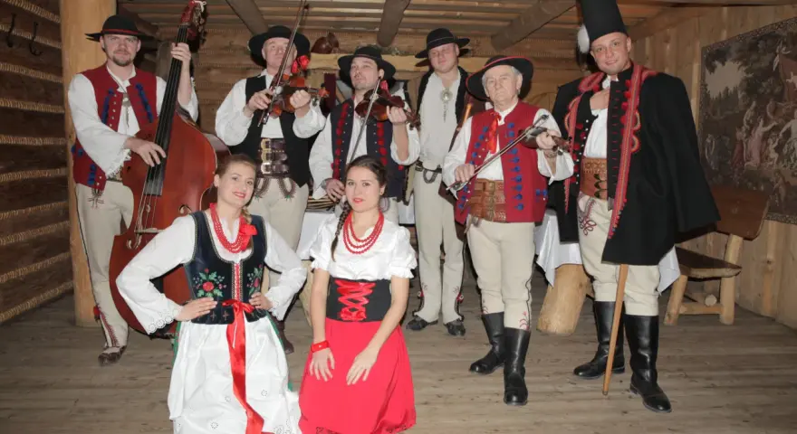 Zespół góralski w strojach ludowych, z instrumentami. Wśród nich skrzypkowie, basista i panie w tradycyjnych sukniach.