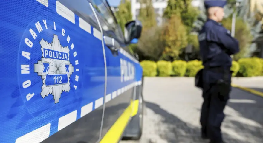 Policjant w mundurze stoi obok radiowozu. Czy zarobki w policji są adekwatne do służby?
