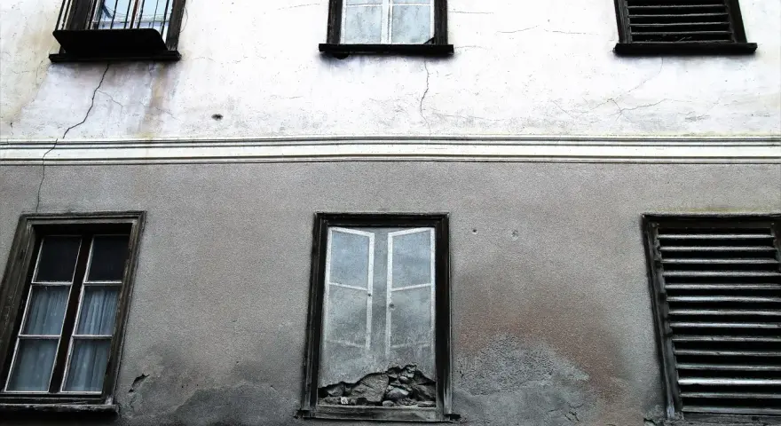Stara elewacja z widocznymi pęknięciami i zaciekami. Okna z żaluzjami i jedno wybite.