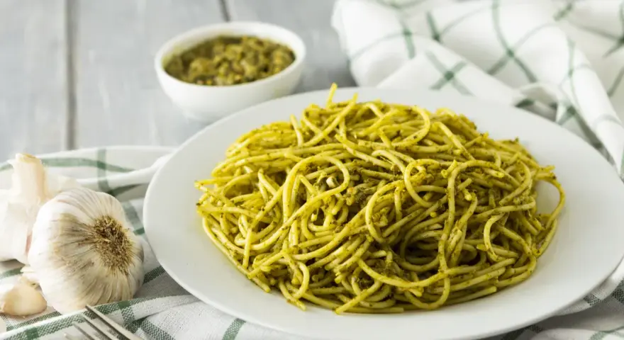 Pyszny makaron z pesto, podany na białym talerzu, obok czosnku i miseczki z sosem.