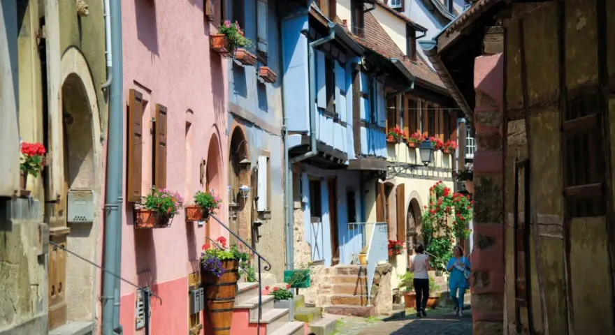 Charming street with colorful houses, perfect for day trips from Strasbourg. Two women stroll past flower-filled window boxes.