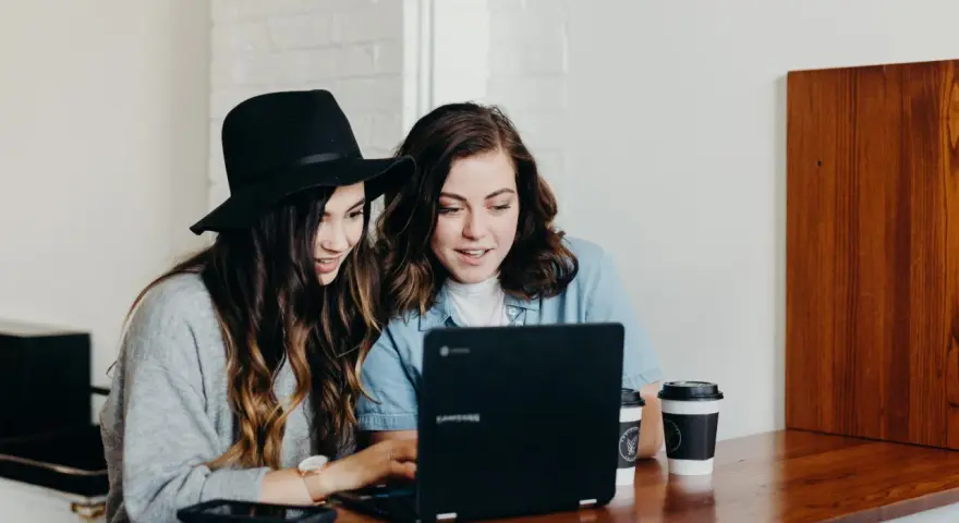 Dwie młode kobiety pracują na laptopie, jedna w kapeluszu, druga w dżinsowej koszuli. Ich wspólny projekt to passe recent.