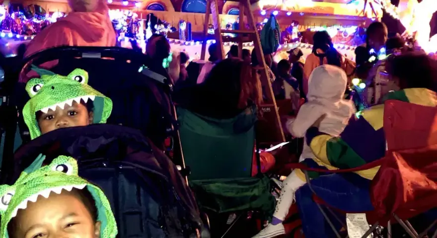 Two kids in dinosaur hoodies watch a vibrant Mardi Gras parade. They're ready for fun at this family-friendly event.