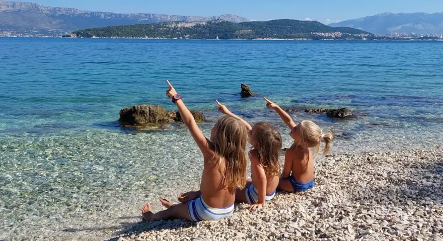 Trzy dziewczynki siedzą na kamienistej plaży w Chorwacji, wskazując palcami na morze. Idealne miejsce, gdzie do Chorwacji z dziećmi.