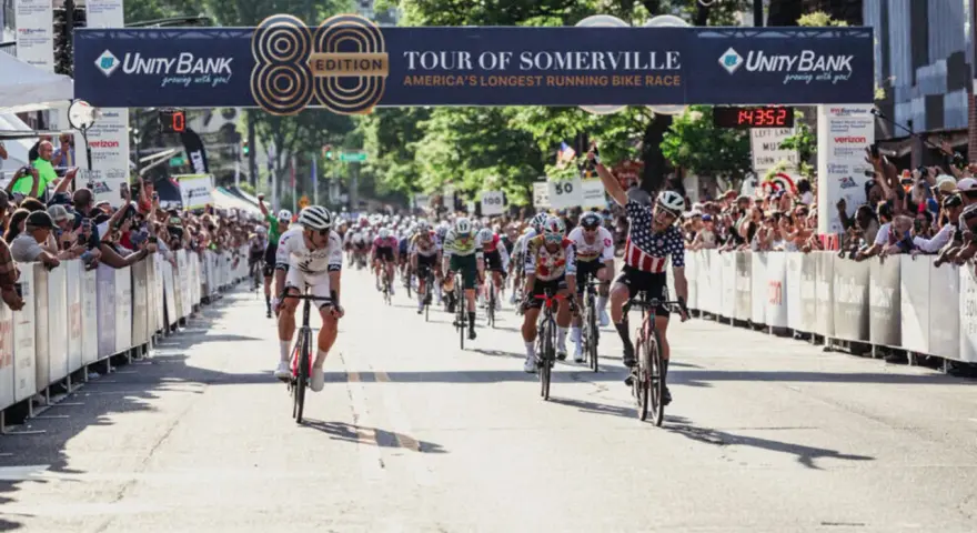 Cyclists race towards the finish line at the Tour of Somerville, America's longest-running bike race, with cheering crowds lining the street.