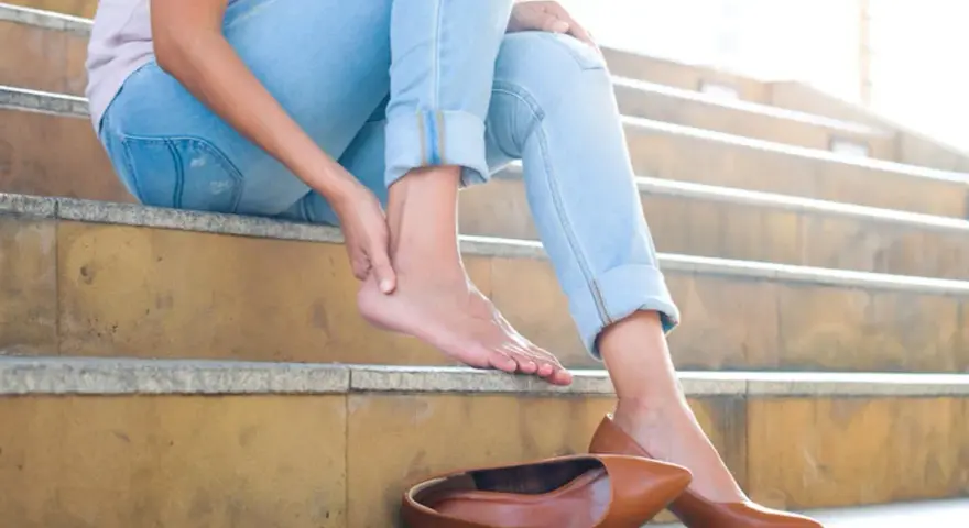 Douleur au talon ? Une femme s'assoit sur des marches, massant son pied. Peut-être un remède de grand-mère pour soulager cette gêne.