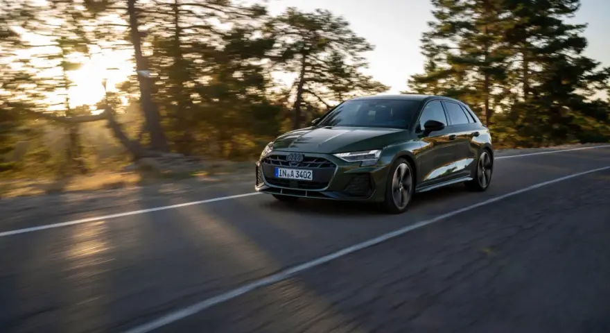 Un'auto verde scuro, un segmento premium, sfreccia su una strada panoramica al tramonto, con alberi sfocati sullo sfondo.