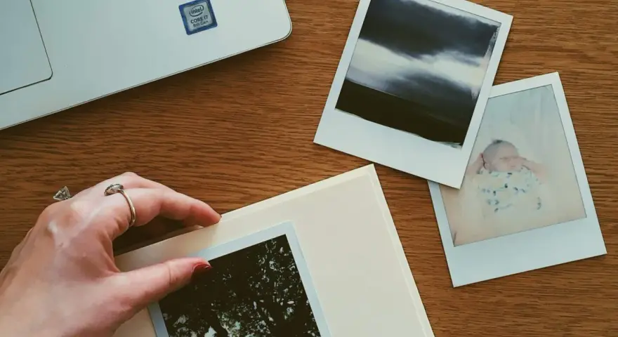 Dłoń układa zdjęcie na albumie, obok inne fotografie i laptop. Idealne miejsce, gdzie przechowywać zdjęcia z telefonu.