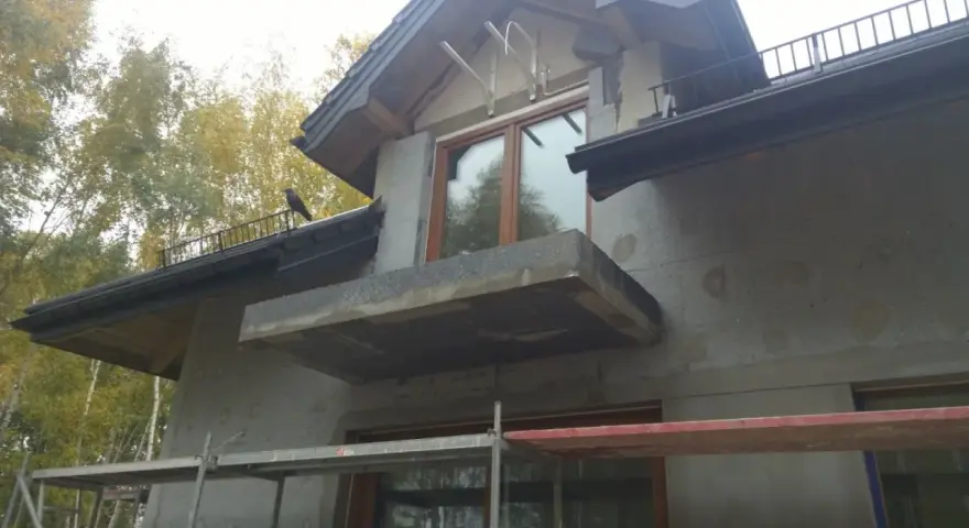 Niedokończona elewacja domu z balkonem i oknem. Na dachu siedzi ptak.