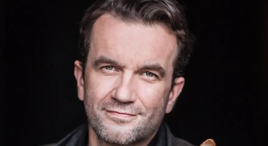 Tomasz Kot zamyślony, z lekkim uśmiechem na twarzy, na czarnym tle.