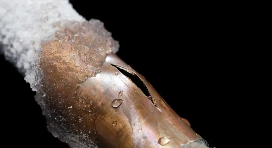 A copper pipe, partially covered in ice, shows a split from a frozen pipe repair. Water droplets cling to the damaged metal.